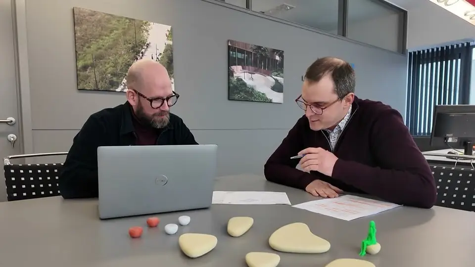 Two men discuss documents at a table with a laptop and materials samples.