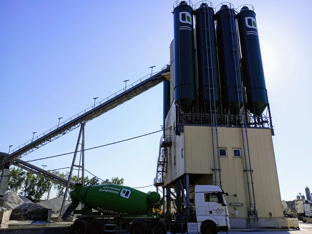 Cementfabriek met vier verticale silo's rechtsboven in beeld en een lange transportband linksboven. Op de voorgrond staat een witte vrachtwagen met een groene cementmixer met bedrijfslogo. Heldere lucht op de achtergrond.